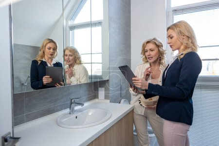 Téléchargez les photos : Agent immobilier ciblé démontrant quelque chose sur la tablette numérique dans la salle de bain à un acheteur potentiel - en image libre de droit