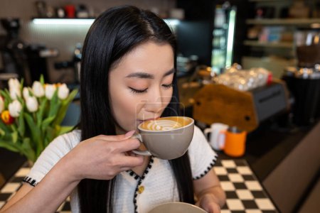 Téléchargez les photos : Portrait d'une brune calme sirotant du cappuccino avec de la mousse d'une tasse en céramique dans le café - en image libre de droit