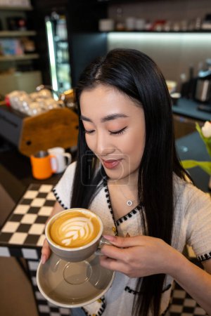 Téléchargez les photos : Souriant contenté attrayant aux cheveux longs asiatique femelle regardant la mousse sur la surface de son cappuccino - en image libre de droit