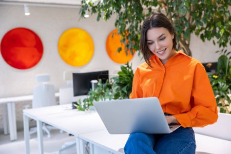 Téléchargez les photos : Souriante jeune femme assise sur le bureau travaillant sur l'ordinateur portable dans ses mains - en image libre de droit