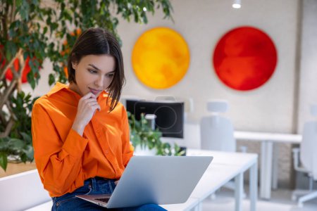 Téléchargez les photos : Tranquille concentré jeune femme caucasienne aux cheveux bruns assis sur le bureau regardant l'écran de l'ordinateur - en image libre de droit