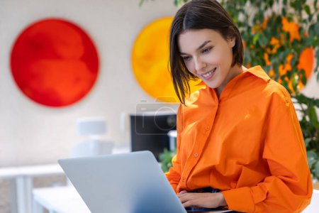 Téléchargez les photos : Souriant contenté belle jeune femme brune assise sur le bureau tapant sur son ordinateur portable - en image libre de droit