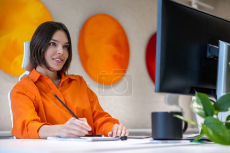 Foto de Empleada concentrada de la empresa sentada en el escritorio de la oficina mirando el monitor de la computadora mientras toma notas en su cuaderno - Imagen libre de derechos