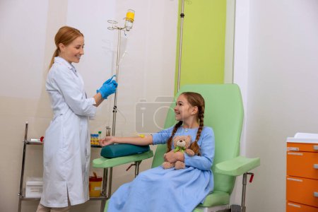 Photo for Little girl kid in clinic room having dropper with medication, doctor with drop counter and patient in hospital sitting in comfortable arm chair. - Royalty Free Image