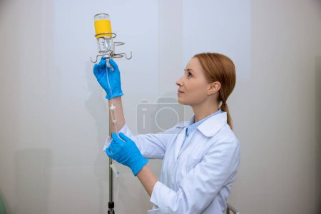 Photo for Blonde young adult doctor or nurse wearing lab coat and gloves with drop counter in hospital. - Royalty Free Image