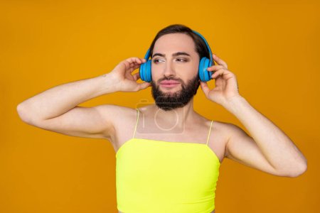 Photo for Dreaming transgender person with beard wearing yellow top posing with headphones listening favorite songs enjoying music isolated over orange background - Royalty Free Image