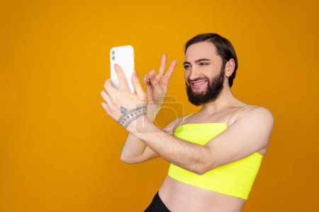 Téléchargez les photos : Souriant satisfait personne transsexuelle barbu portant haut jaune posant selfie montrant v signe morceau geste isolé sur fond orange - en image libre de droit