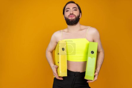 Téléchargez les photos : Homme d'affaires transsexuel barbu confiant portant des dossiers de tenue supérieure jaune regardant la caméra faire des travaux de papier posant isolé sur fond orange - en image libre de droit