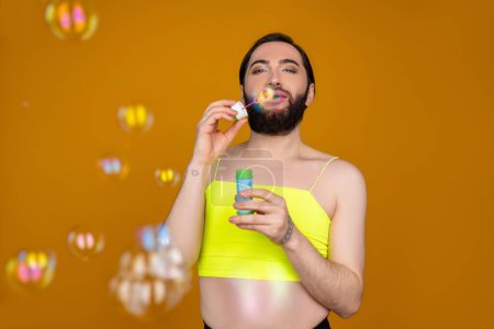 Téléchargez les photos : Ludique personne transsexuelle barbu portant haut jaune soufflant des bulles de savon s'amusant posant isolé sur fond orange - en image libre de droit