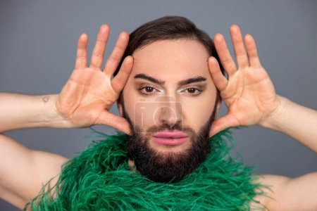 Foto de Serio moreno transexual persona con maquillaje usando boa verde bufanda mantiene las palmas cerca de la cara mirando la cámara posando aislado sobre fondo gris - Imagen libre de derechos