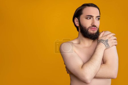 Téléchargez les photos : Pensif rêvant personne transgenre nue avec barbe cachant son corps, détournant les yeux, copier l'espace pour le texte promotionnel, posant isolé sur fond orange - en image libre de droit