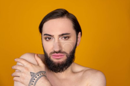 Téléchargez les photos : Gros plan portrait de la personne transgenre nue brune avec barbe posant avec les épaules nues en regardant la caméra isolée sur fond orange - en image libre de droit