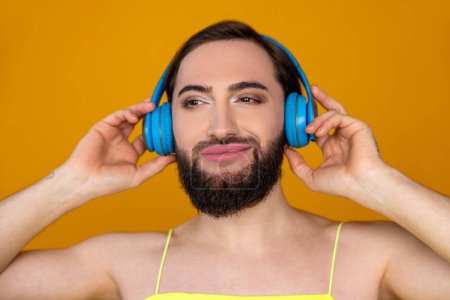 Téléchargez les photos : Portrait rapproché d'une personne transgenre satisfaite et souriante avec barbe écoutant de la musique via des écouteurs bleus prenant plaisir à poser isolée sur fond orange - en image libre de droit