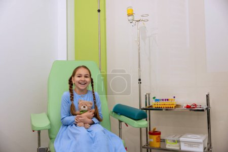 Téléchargez les photos : Petite fille enfant dans la chambre d'hôpital ayant compte-gouttes avec des médicaments, assis dans un fauteuil confortable. - en image libre de droit