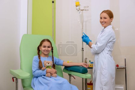 Téléchargez les photos : Petite fille enfant dans la salle de clinique ayant compte-gouttes avec des médicaments, médecin avec comptoir de chute et patient à l'hôpital assis dans un fauteuil confortable. - en image libre de droit