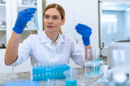 Téléchargez les photos : Femme blonde chercheuse à succès travaillant avec des échantillons médicaux, tenant un tube à essai avec un liquide bleu, faisant des recherches en microbiologie en laboratoire. - en image libre de droit