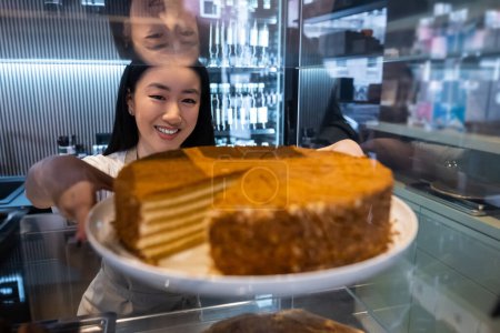 Téléchargez les photos : Travailleur de café joyeux prenant un plateau de service avec un dessert savoureux de l'étagère vitrine de la pâtisserie en verre - en image libre de droit