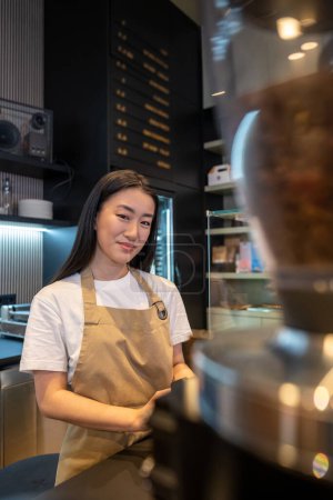 Foto de Sonriendo complacido lindo joven asiático empleado de pie en el mostrador en un café moderno - Imagen libre de derechos