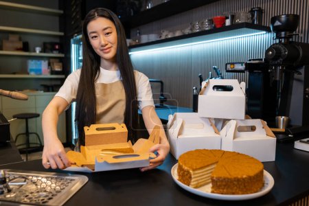 Téléchargez les photos : Travailleur de café debout au comptoir tout en emballant une tranche de gâteau dans la boîte en carton - en image libre de droit