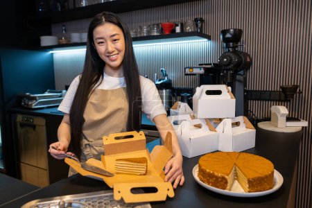 Photo for Smiling happy cute young Asian cafe worker placing a slice of cake into the box - Royalty Free Image