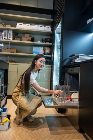 Photo for Smiling young coffee shop employee sitting on her haunches in front of the open dishwasher filled with dishware - Royalty Free Image