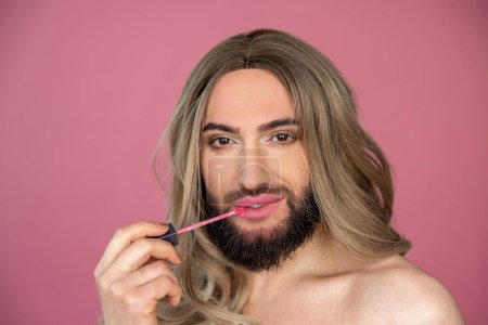 Photo for Beauty portrait of transgender wearing blonde wig and white dress doing makeup applying lip gloss preparing for party posing isolated over pink background - Royalty Free Image