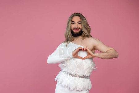 Photo for Smiling cheerful transgender wearing blonde wig and white dress making heart shape with hands, expressing love, posing isolated over pink background - Royalty Free Image