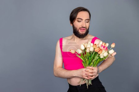 Téléchargez les photos : Attrayant romantique brunette transsexuelle portant haut rose tenant bouquet de fleurs de printemps admire la beauté posant isolé sur fond gris avec espace de copie - en image libre de droit