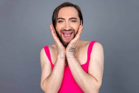 Téléchargez les photos : Positif heureux gai brunette transsexuelle portant haut rose regardant la caméra avec sourire touchant visage posant isolé sur fond gris - en image libre de droit
