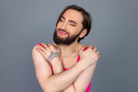 Téléchargez les photos : Satisfait auto aimant brunette transsexuelle portant haut rose s'embrassant avec le sourire et le plaisir posant isolé sur fond gris - en image libre de droit