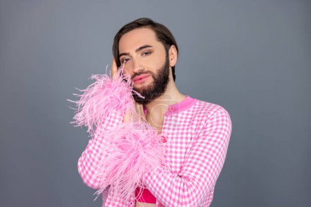 Téléchargez les photos : Mignon souriant transgenre personne portant des vêtements roses appuyé sur les mains en regardant la caméra posant isolé sur fond gris - en image libre de droit