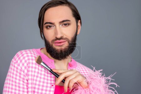 Téléchargez les photos : Beau attrayant grave transgenre personne portant des vêtements roses faire maquillage, tenant brosse visage dans les mains posant isolé sur fond gris - en image libre de droit