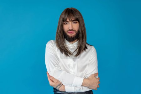 Photo for Attractive transgender person wearing brunette wig and white shirt hugging himself protects personal individuality posing isolated over blue background - Royalty Free Image