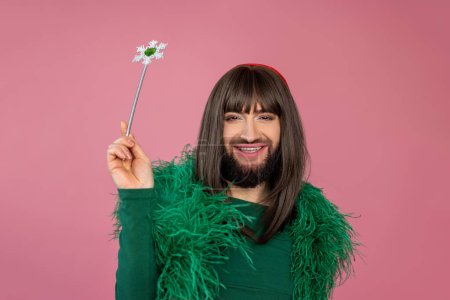 Téléchargez les photos : Joyeux transgenre souriant avec maquillage et perruque portant une robe de soirée en plumes vertes jouant avec baguette magique posant isolé sur fond rose - en image libre de droit