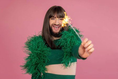 Téléchargez les photos : Ravi de rire transgenre avec maquillage et perruque portant une robe de soirée en plume verte tenant scintillant hurlant d'excitation posant isolé sur fond rose - en image libre de droit