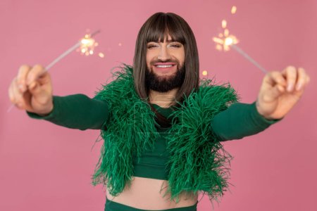Photo for Joyful transsexual with makeup and wig wearing green feather dress having fun with sparklers, spending leisure time on party posing isolated over pink background - Royalty Free Image