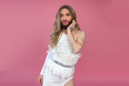 Foto de Confiado atractivo transexual vistiendo peluca rubia y vestido blanco de pie tocando la cara mirando a la cámara posando aislado sobre fondo rosa - Imagen libre de derechos