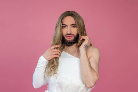 Photo for Beautiful transgender wearing blonde wig and white dress looking at camera with confident expression posing isolated over pink background - Royalty Free Image