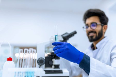 Foto de Sonriente doctor trabajando en moderno laboratorio investigador científico examina sustancia bajo el microscopio, sosteniendo tubo de ensayo con líquido azul en las manos. - Imagen libre de derechos
