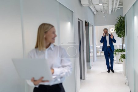 Téléchargez les photos : Homme d'affaires joyeux appelant sur le smartphone tout en marchant vers sa collègue le long du couloir du bureau - en image libre de droit