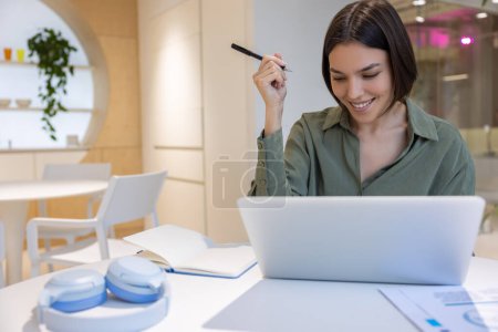 Téléchargez les photos : Travailleur d'entreprise souriant avec un stylo dans la main assis au bureau en regardant l'écran de l'ordinateur portable - en image libre de droit
