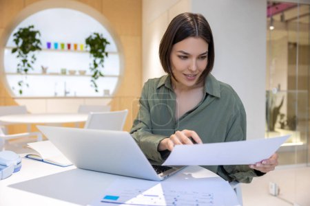 Téléchargez les photos : Une employée d'entreprise concentrée assise à l'ordinateur portable regardant un document entre ses mains - en image libre de droit