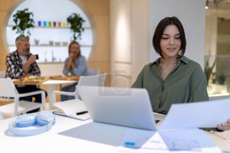 Photo for Smiling corporate worker sitting at the portable computer while looking at a document in her hand - Royalty Free Image