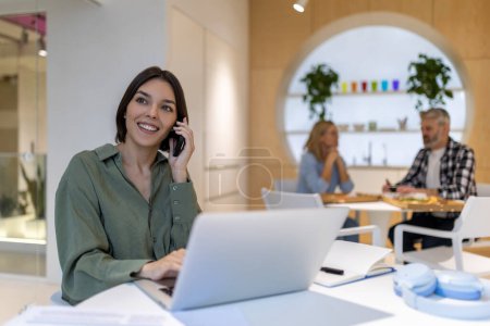 Photo for Smiling pleased young female company employee sitting at the portable computer while calling on the cellphone - Royalty Free Image