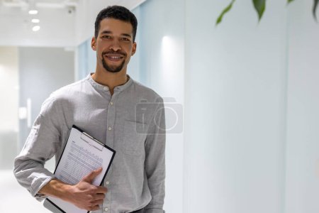 Photo for Waist-up portrait of a joyous corporate employee with the laptop and business documentation in the hand standing in the corridor - Royalty Free Image