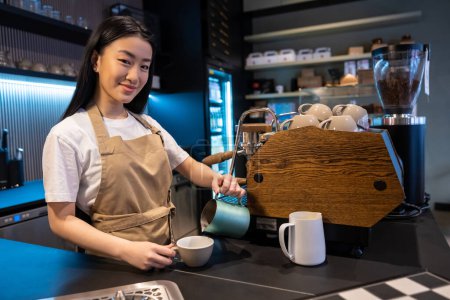 Téléchargez les photos : Souriant amical jeune asiatique barista verser une boisson de la cruche dans la tasse vide - en image libre de droit
