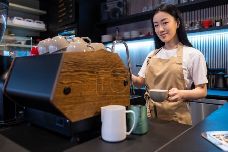 Téléchargez les photos : Souriant employé asiatique café debout à la machine à café avec une tasse en céramique dans la main - en image libre de droit
