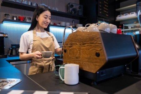 Foto de Concentrado barista lindo de pie en la máquina de café con una taza de cerámica en la mano - Imagen libre de derechos