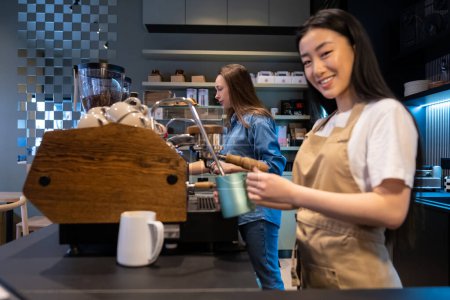 Téléchargez les photos : Joyeux jeune travailleur de café debout à la machine à café avec une cruche de lait dans la main à côté de son collègue sérieux - en image libre de droit