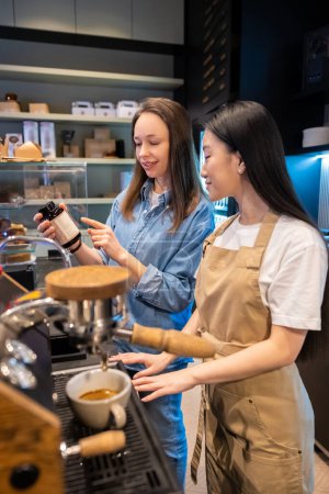 Photo for Barista looking at a plastic bottle in her female colleague hands while making a caffeinated beverage with the coffee machine - Royalty Free Image